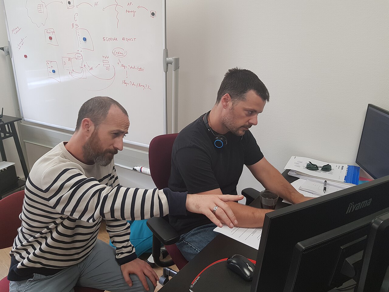 Two software developers at a desk reviewing code together on a monitor, with a whiteboard architecture sketch behind them
