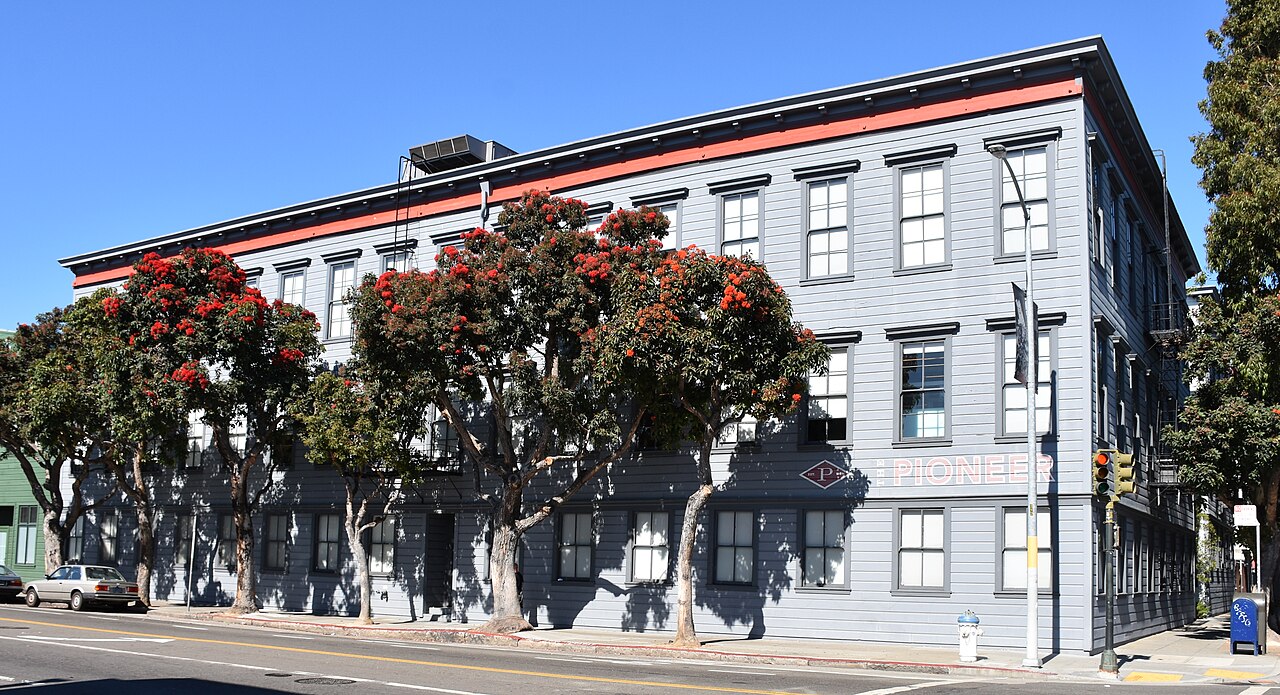 Three-storey grey wooden Pioneer Building with red trim on a San Francisco street corner, with flowering trees in front
