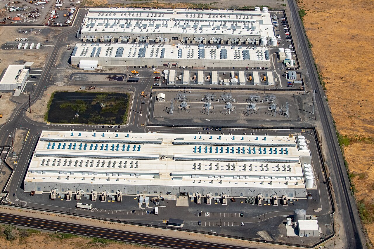 Aerial photograph of an Amazon Web Services data center campus with large rooftop cooling equipment, an electrical substation and a pond