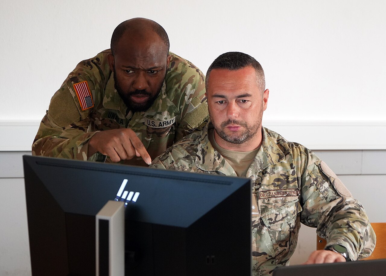 A US Army National Guard cyber analyst pointing at a monitor while a partner-nation officer works at a computer during cyber skills training