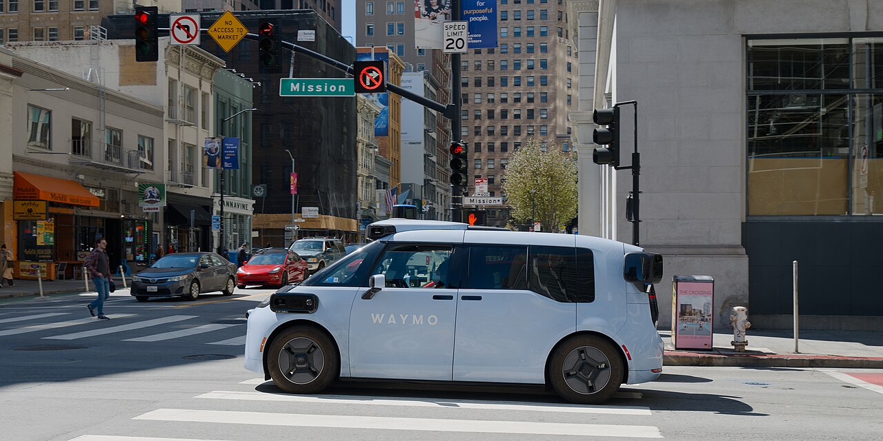 A white Waymo Ojai robotaxi stopped at an intersection on Mission Street in San Francisco, with traffic signals and a crosswalk in view