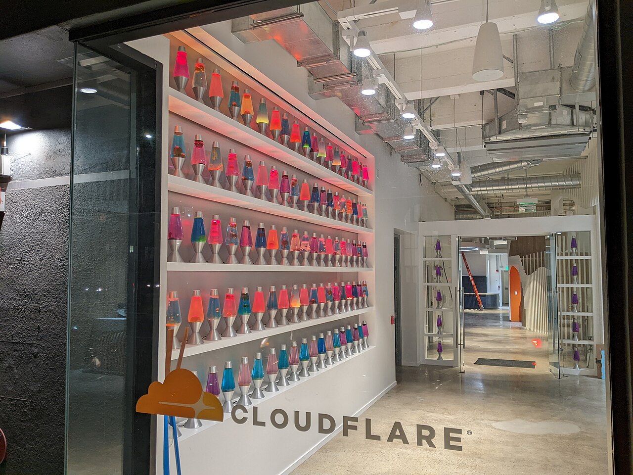 A wall of colorful lava lamps behind glass at Cloudflare's San Francisco office, with the Cloudflare logo visible on the window
