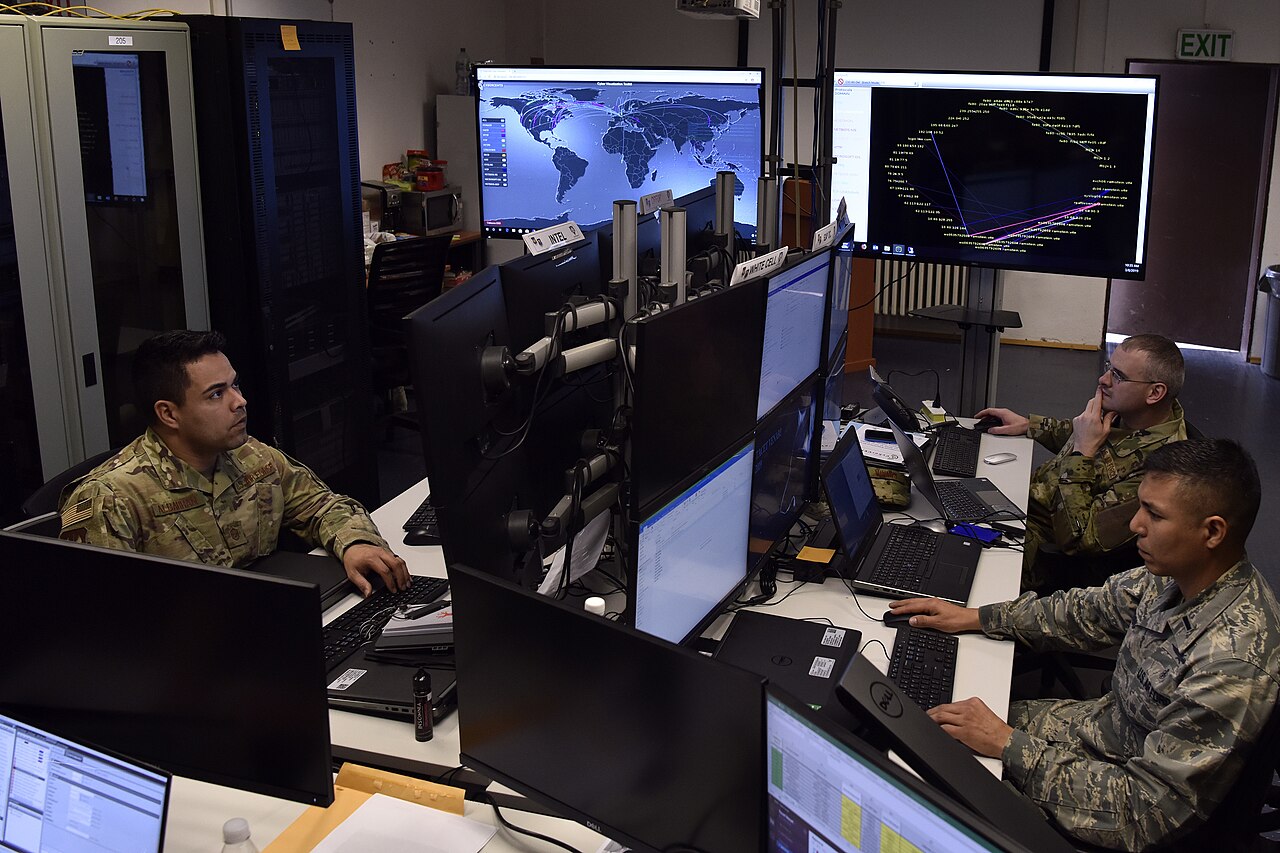 U.S. Air Force cyber exercise support staff working at multi-monitor consoles with network and attack-path visualizations during a defensive cyber operations training exercise
