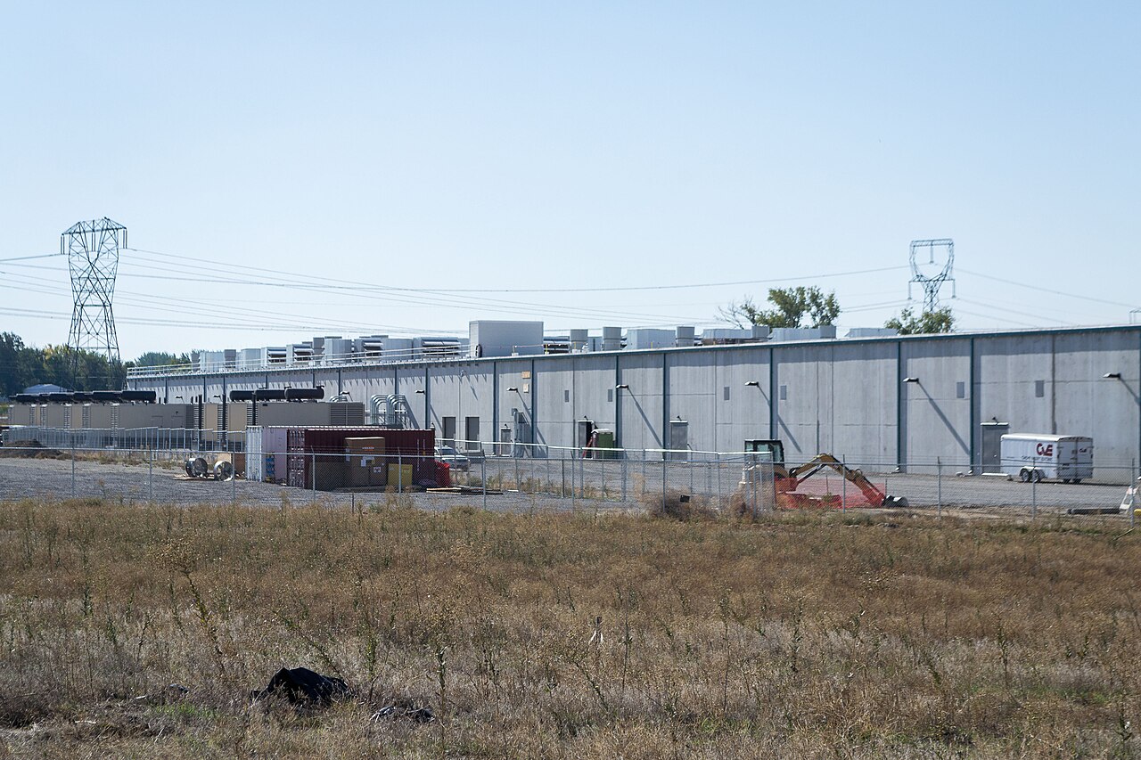 Long, low grey Amazon data center building in Boardman, Oregon, behind a fence with power transmission towers