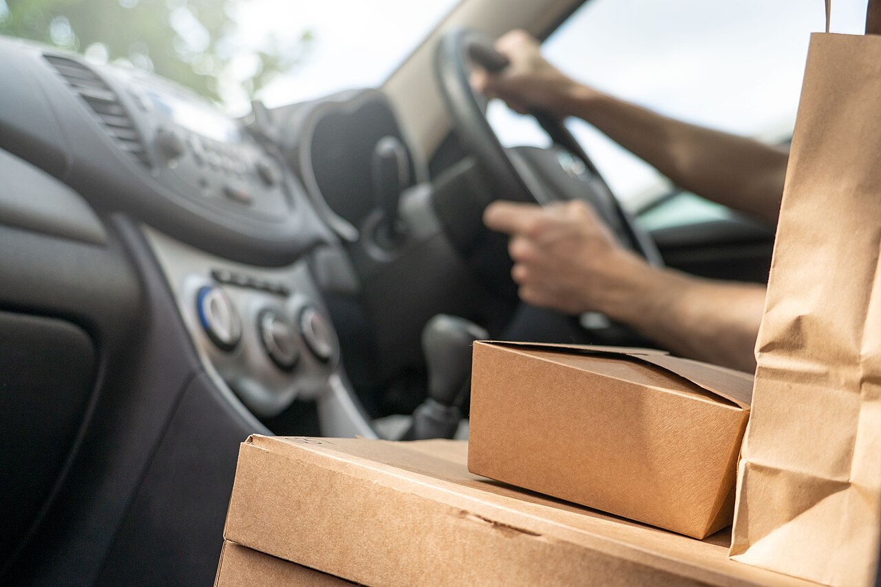 A delivery driver's hands on a steering wheel beside takeaway boxes and a paper bag on the passenger seat