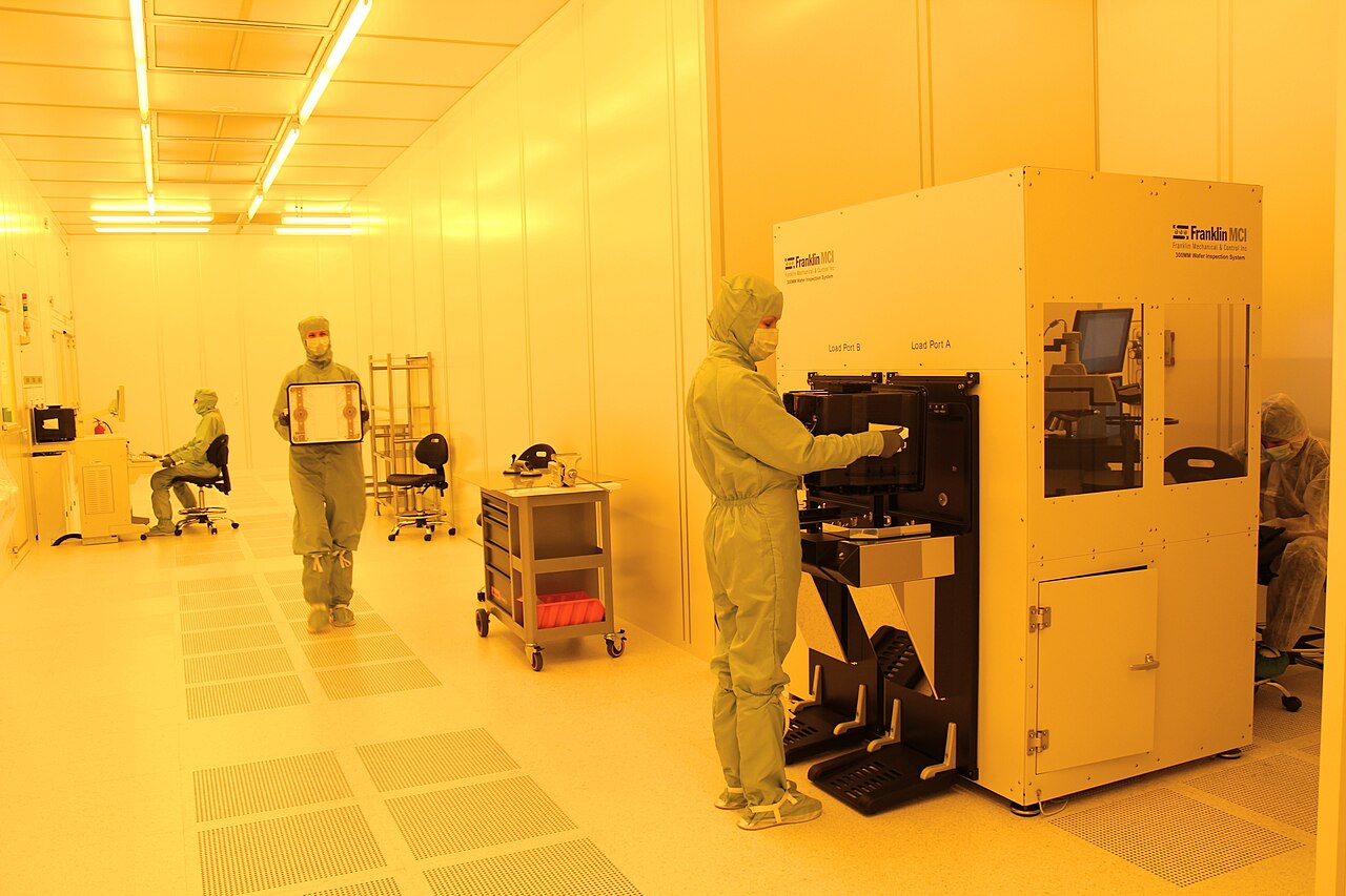 Technicians in cleanroom suits working beside a 300mm wafer inspection system in a semiconductor fab