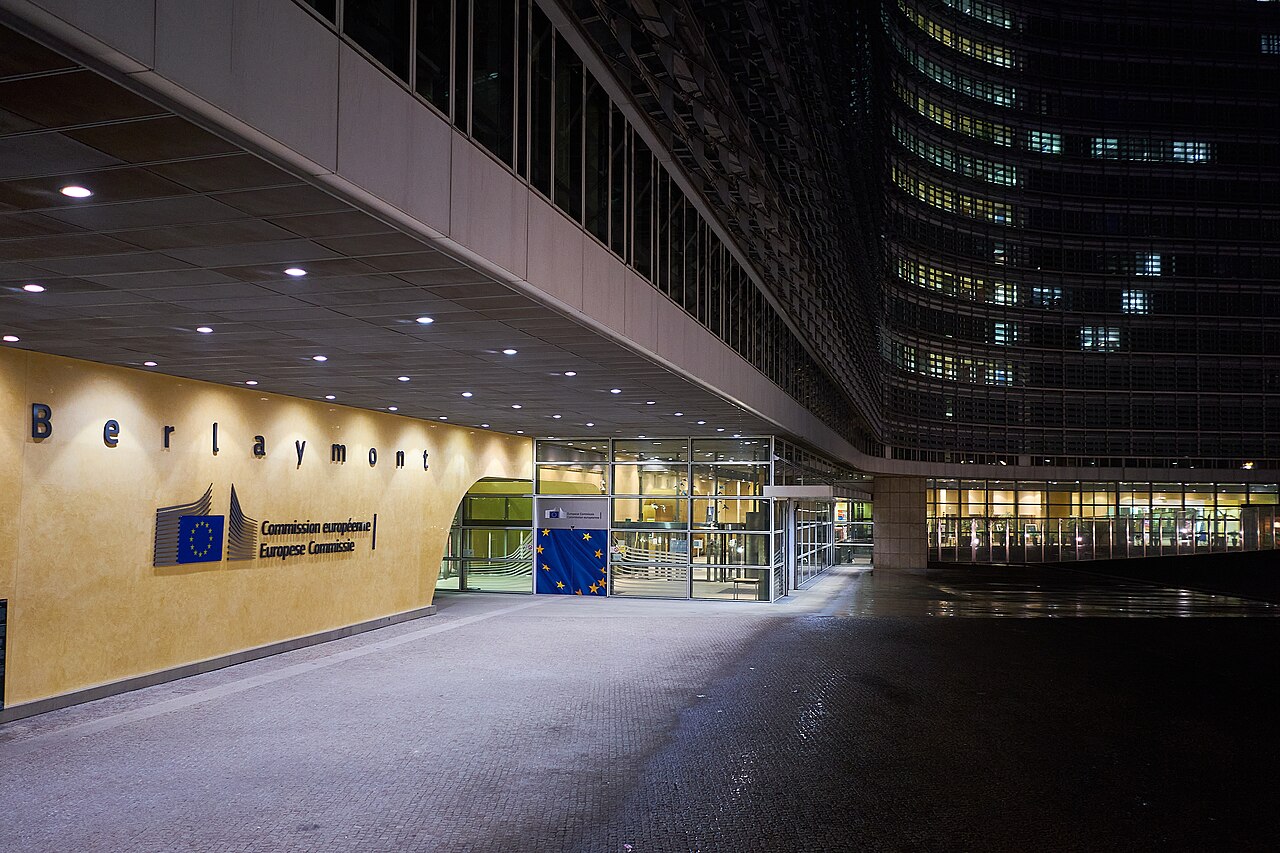 The entrance of the Berlaymont building at night, with the European Commission name and logo lit above a European Union flag