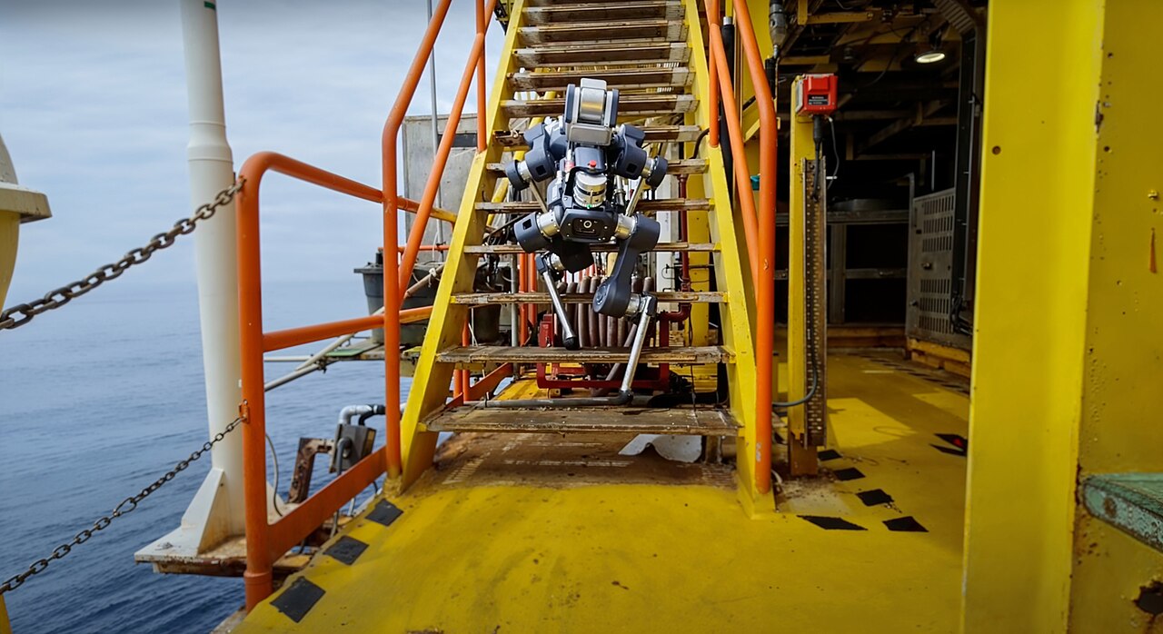 An ANYmal quadruped robot climbing a yellow steel staircase on an offshore platform, with the sea visible to the left