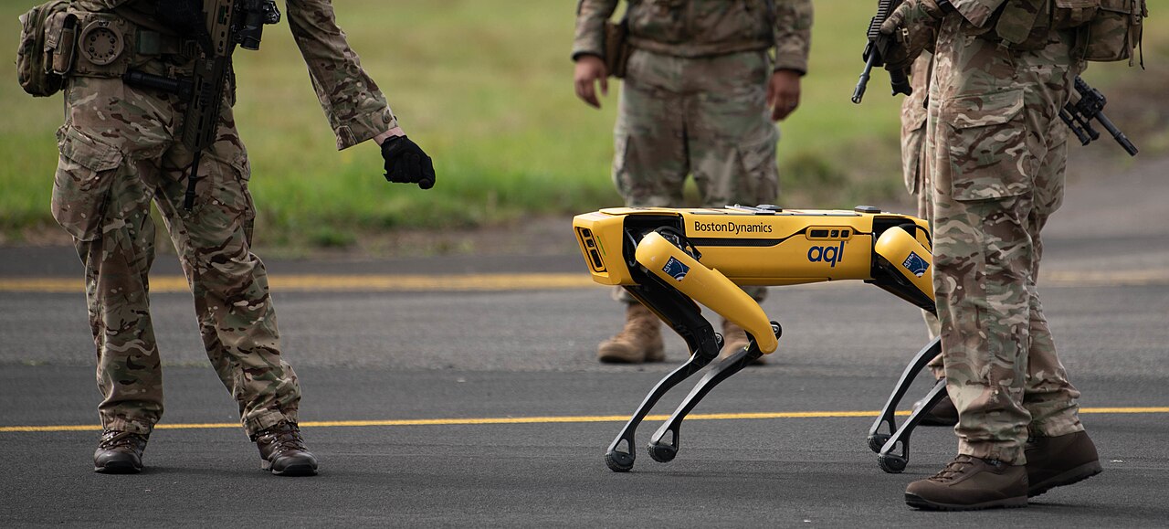 A yellow Boston Dynamics Spot quadruped robot walking on an airfield among Royal Air Force personnel in camouflage