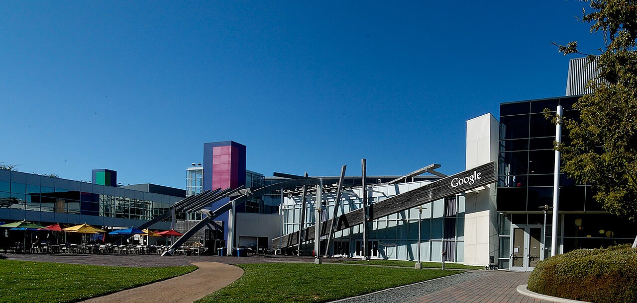 Google campus buildings with the Google name on a wooden fascia and a courtyard with colored umbrellas in front