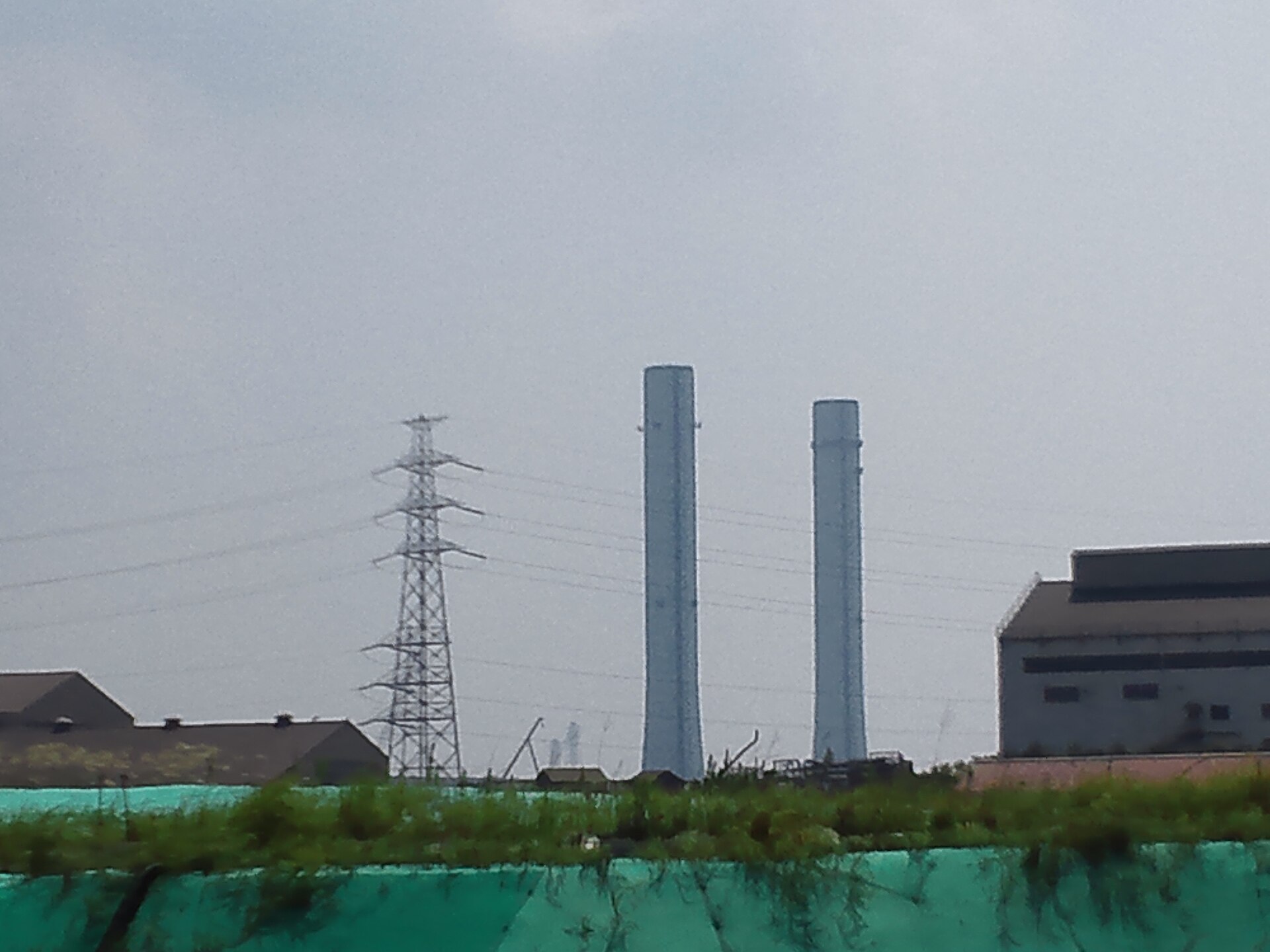 Two tall white exhaust stacks and a transmission tower at the Chiba Thermal Power Station seen from outside the site under an overcast sky