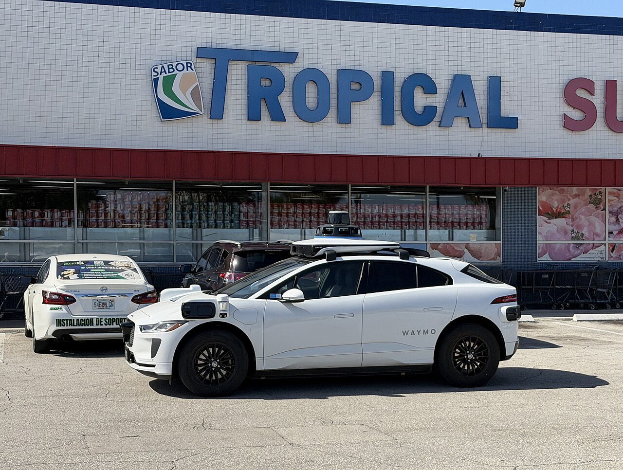 A white Waymo Jaguar I-PACE robotaxi with roof and fender sensor pods parked in a supermarket lot