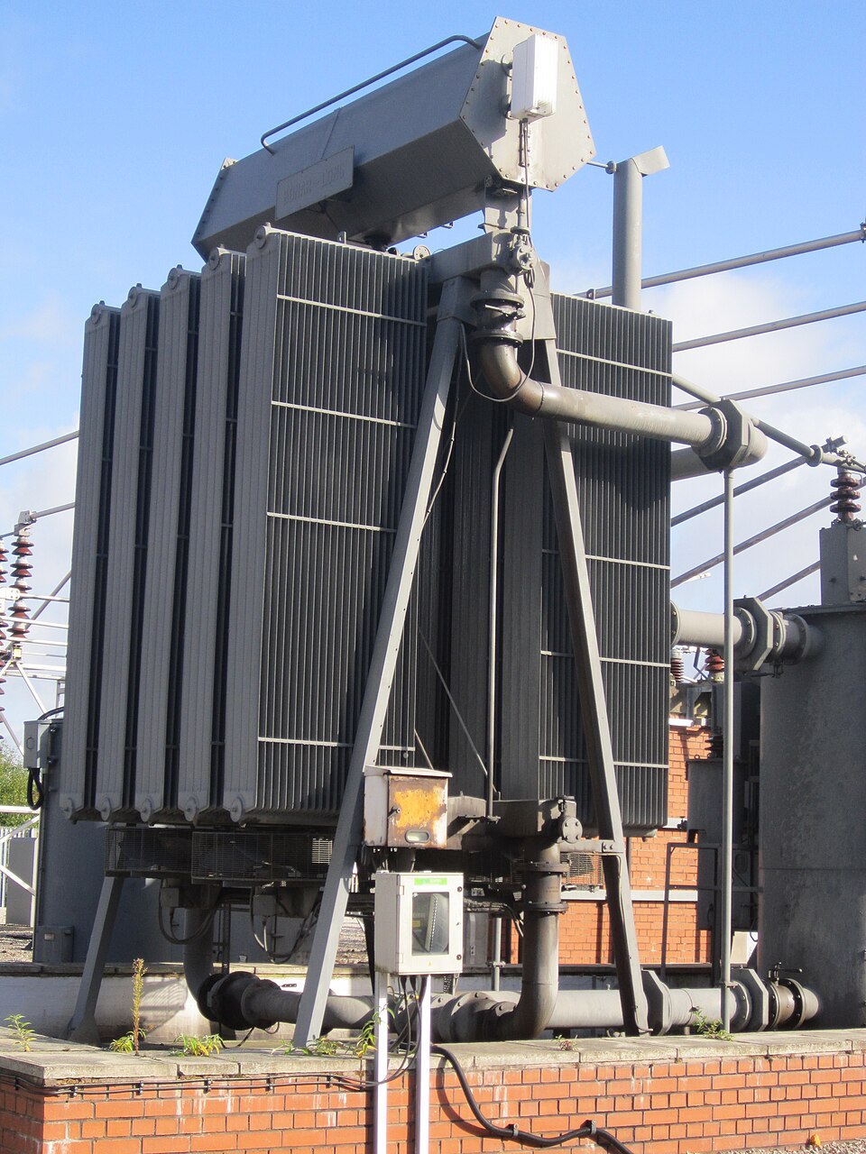A large grey electrical power transformer with cooling radiator fins and insulators at an outdoor substation under a blue sky