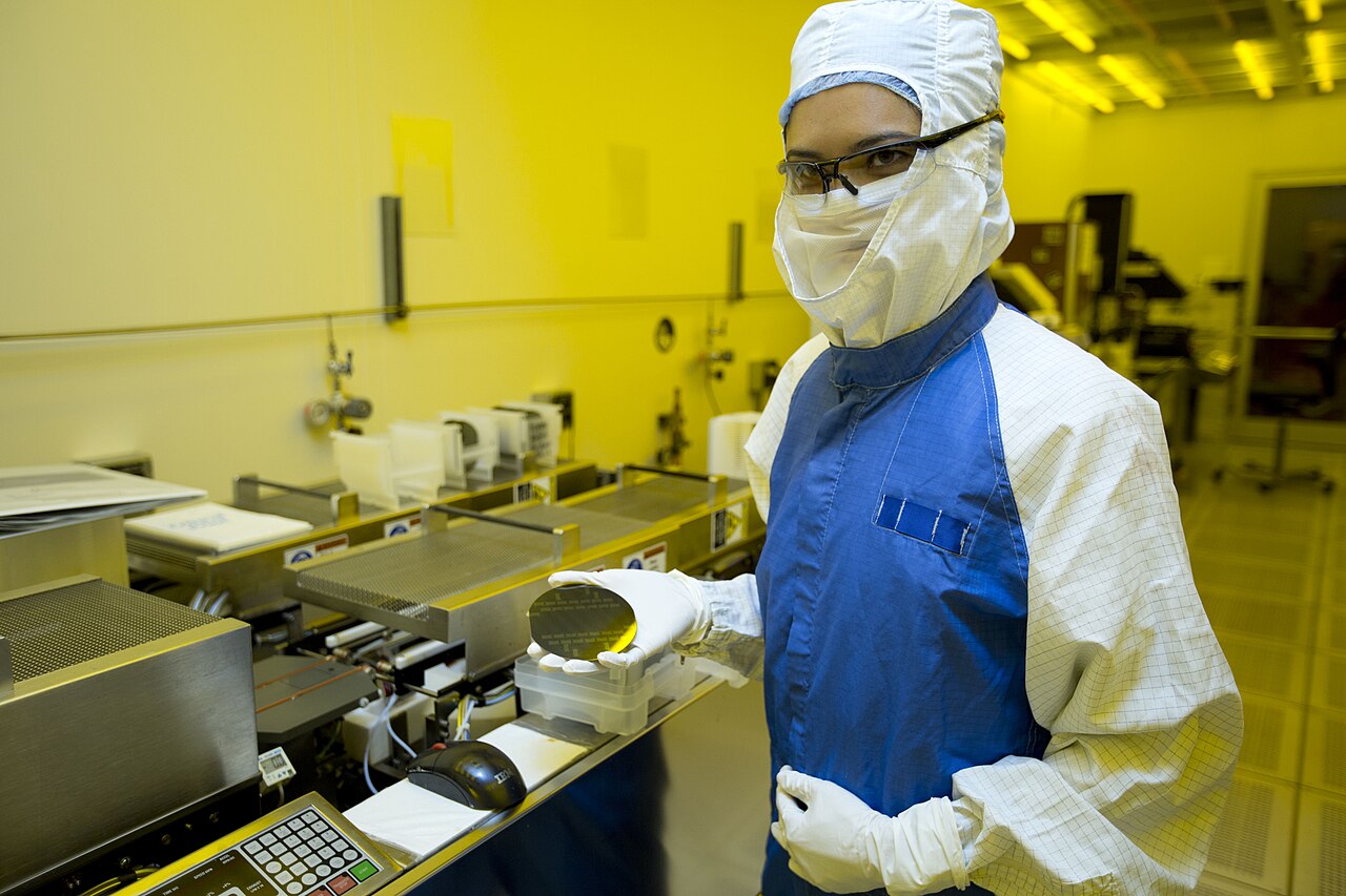 A researcher in cleanroom protective gear holding a silicon wafer inside a semiconductor fabrication lab lit in yellow light