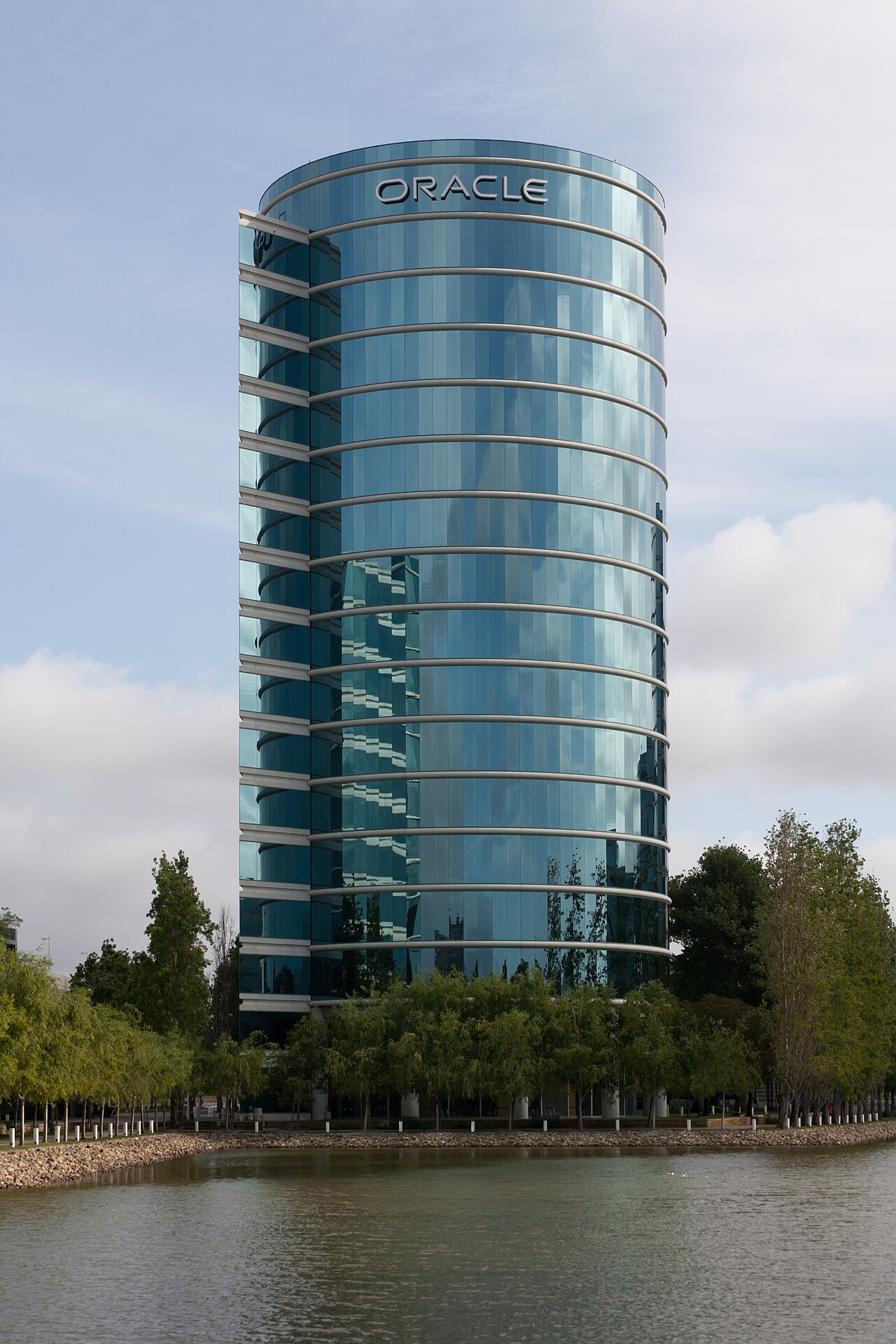 A cylindrical glass Oracle headquarters tower with the ORACLE name visible near the roofline, photographed from across the Redwood Shores lagoon