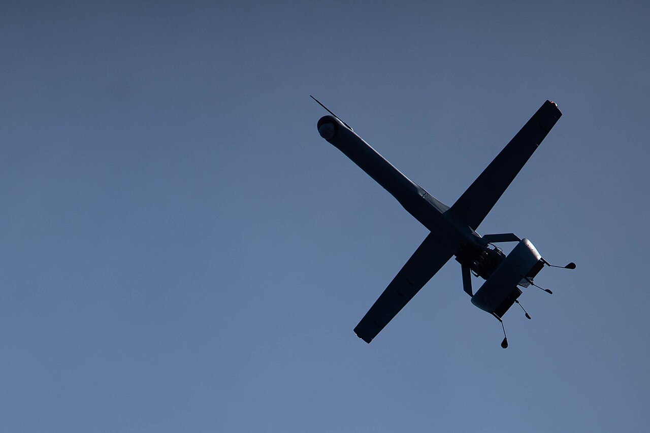 A Shield AI V-BAT drone, seen from below in its distinctive X-wing tail-sitter configuration, flying above the amphibious ship USS Rushmore