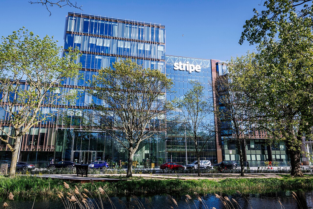 The glass facade of Stripe's Dublin office building with the lowercase stripe wordmark on the front, framed by trees and a canal in the foreground