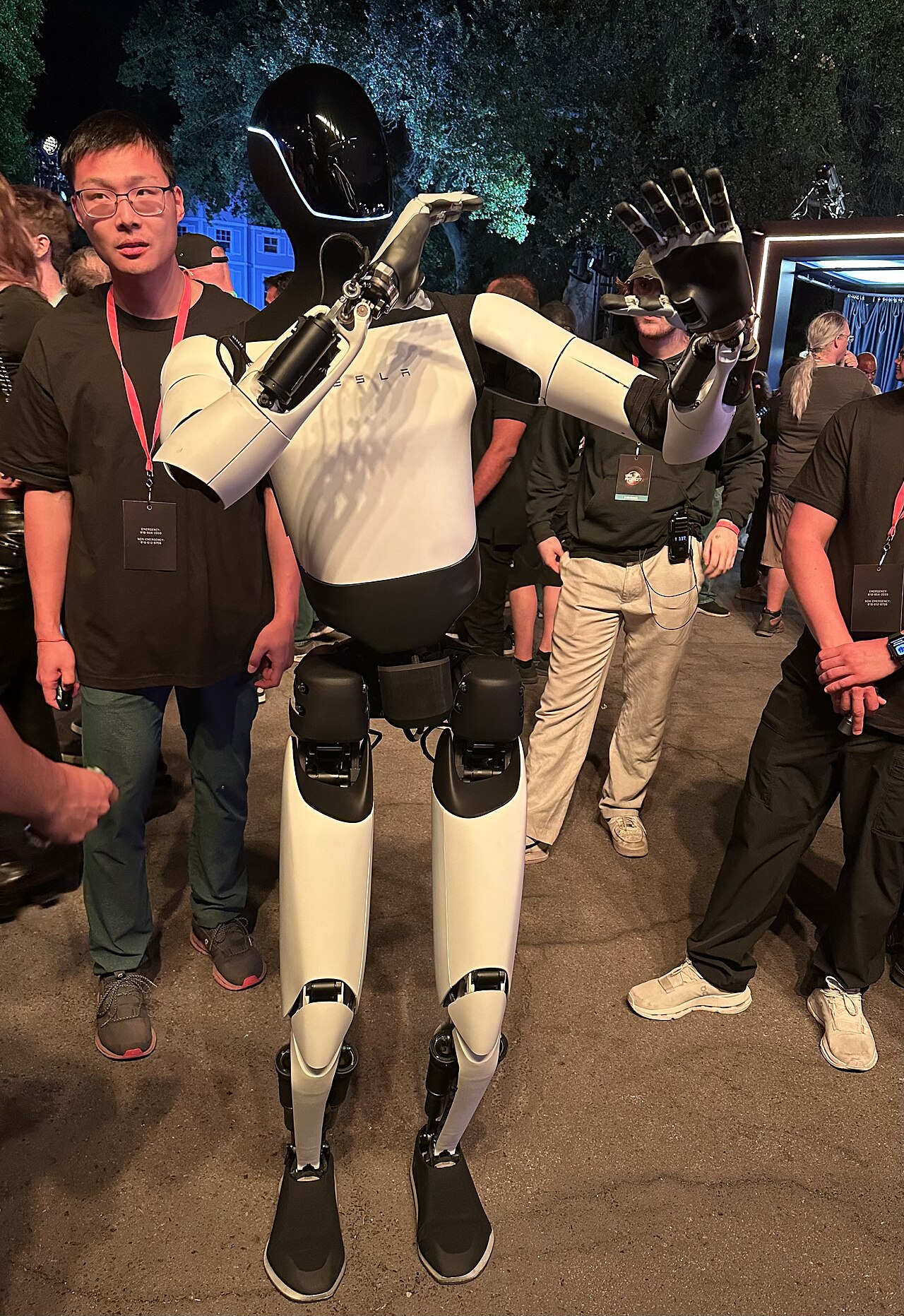 A Tesla Optimus humanoid robot standing among people at an event, with its articulated hands visible