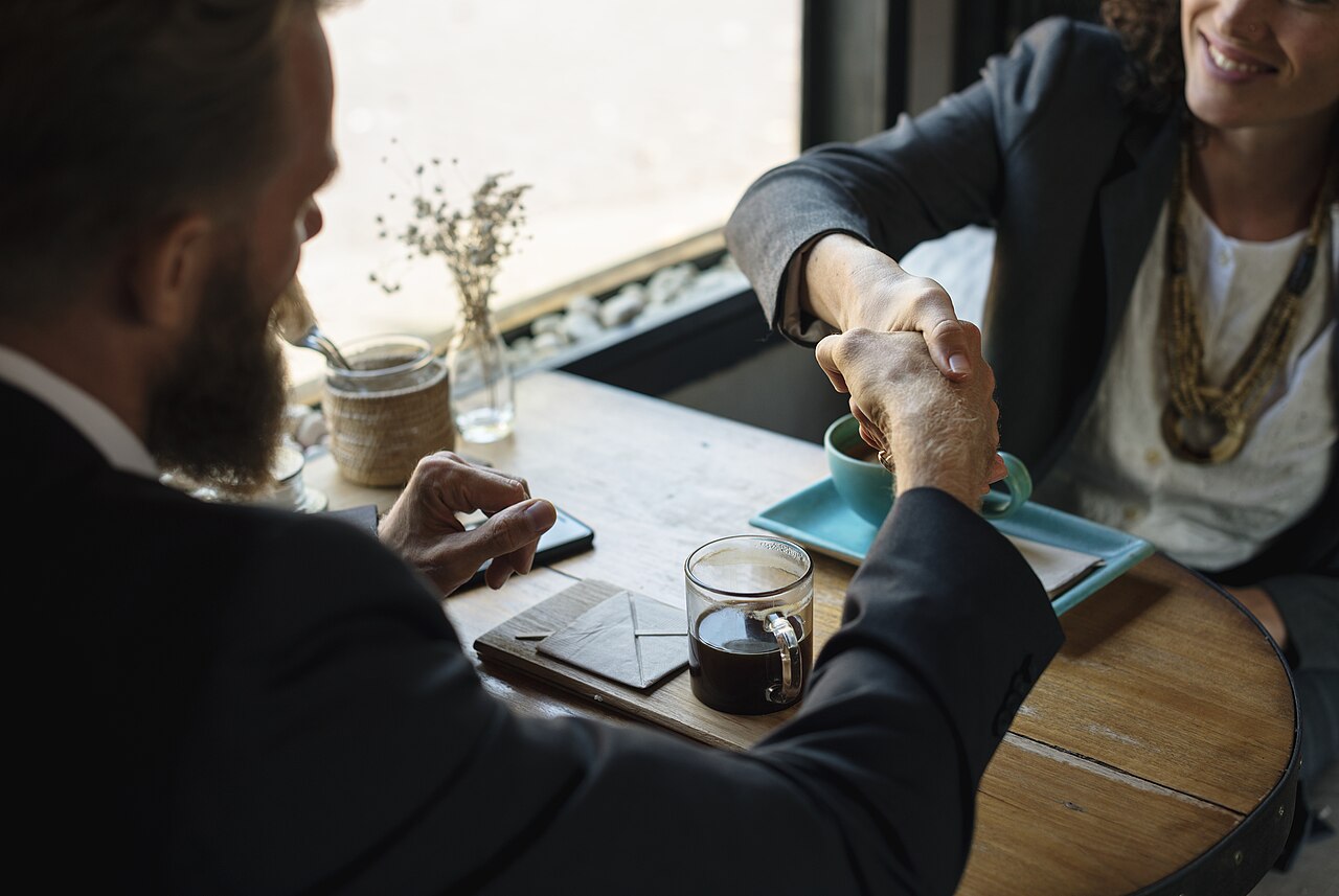 Two people shaking hands over a business agreement at a table, illustrative of a hiring or onboarding moment