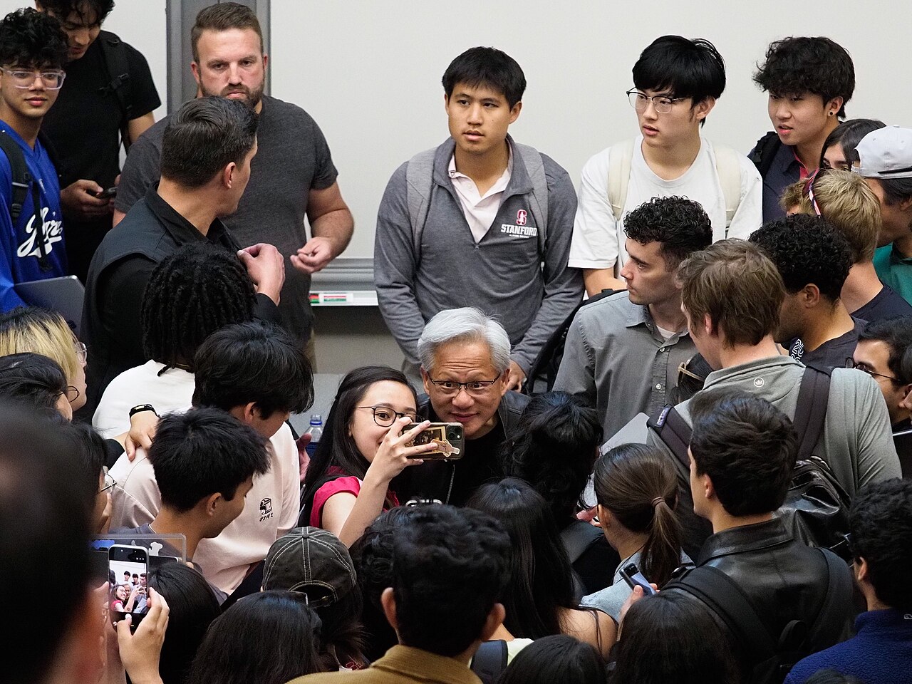 Nvidia CEO Jensen Huang surrounded by a crowd of students at Stanford, several taking photos