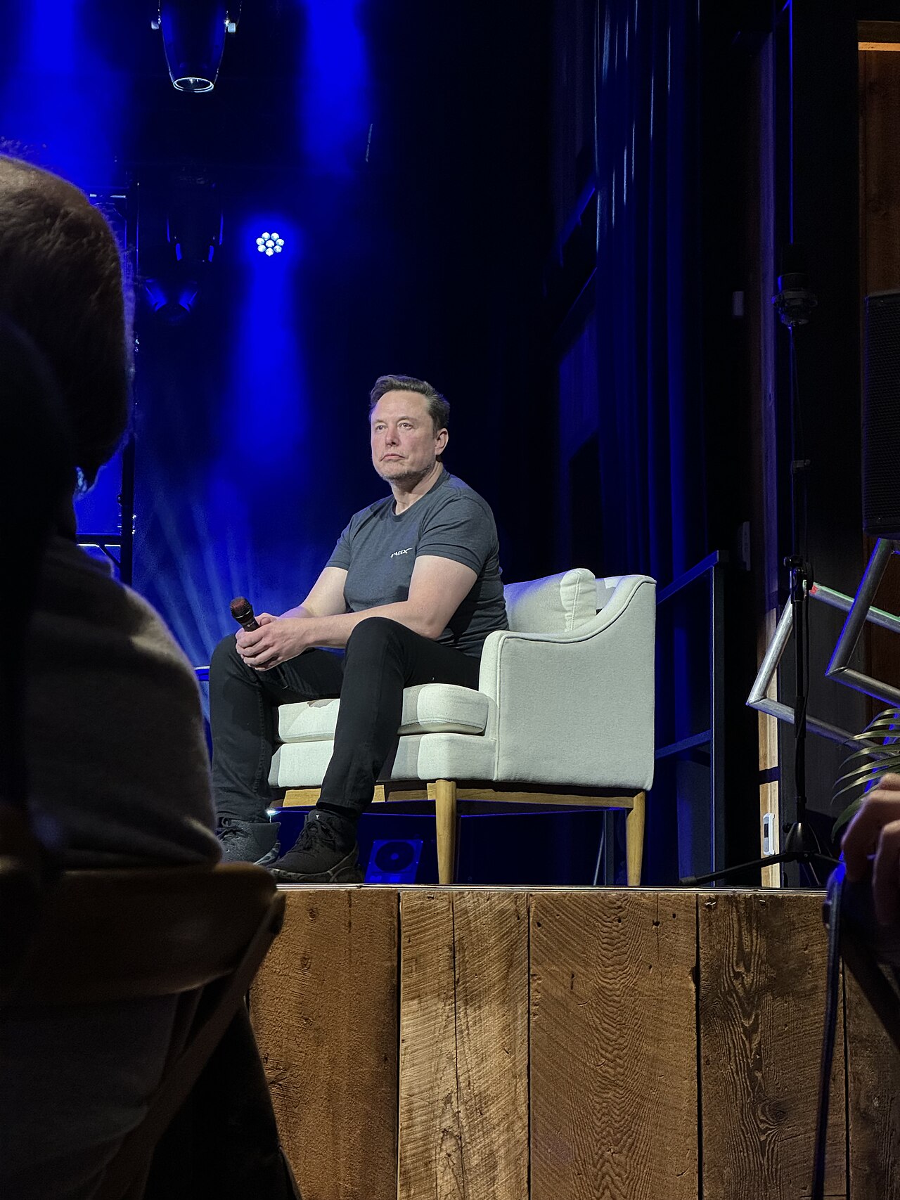 Elon Musk seated on a stage holding a microphone, lit in blue light, during a public conversation